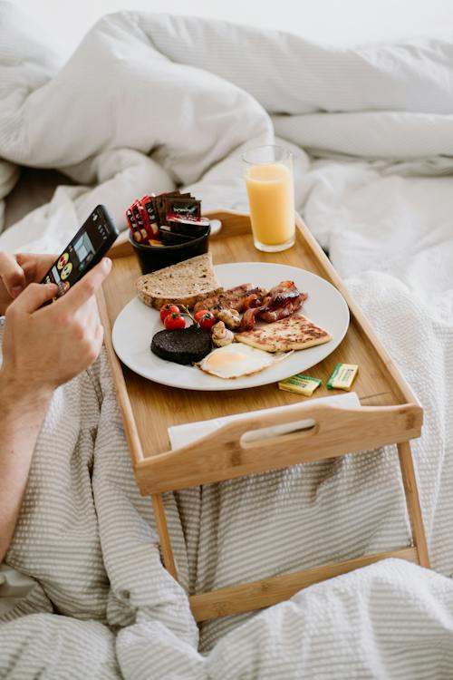 Crop person with smartphone and delicious breakfast in bed